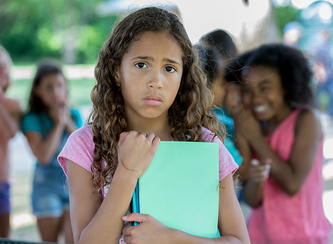 a girl with long wavy hair holds binders to her chest with a sad face while peers behind her appear to be laughing