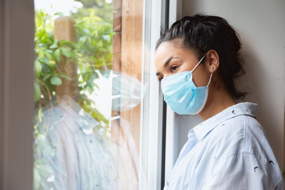 Young hispanic woman, wearing protective face mask, standing by the window and looking out to the street in times of coronavirus crisis, staying at home